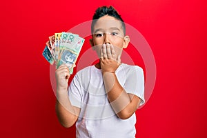 Little boy hispanic kid holding australian dollars covering mouth with hand, shocked and afraid for mistake