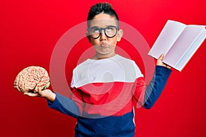 Little boy hispanic kid holding brain studying for school making fish face with mouth and squinting eyes, crazy and comical