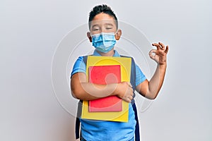 Little boy hispanic kid wearing medical mask going to school doing ok sign with fingers, smiling friendly gesturing excellent