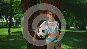 Little boy hold soccerball lean on tree. Young athlete posing with ball closeup.