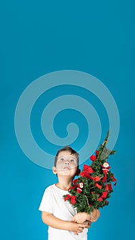 Little boy holding a Christmas tree in his hand and looking up, advertising space, blue backround