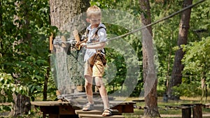 Little boy holding safety rope and walking over wobbly wooden bridge at adventure park. Active childhood, healthy lifestyle, kids