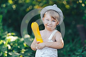 Little boy holding yellow zucchini squash in summer garden farmer boy in white casual robe and hat