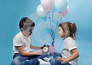 Little boy holds an alarm clock and shows it to his sister on the background of colored balloons