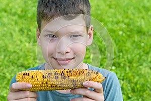 Little boy holds in his hands a fried yellow ear of corn and wants to eat it, sitting on the green grass