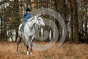 Little boy in a jockey cap on a white adult horse on a background of nature. Jockey, hippodrome, horseback riding