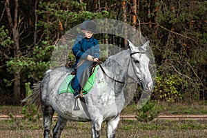 Little boy in a jockey cap on a white adult horse on a background of nature. Jockey, hippodrome, horseback riding