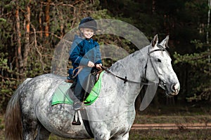 Little boy in a jockey cap on a white adult horse on a background of nature. Jockey, hippodrome, horseback riding