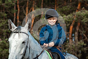 Little boy in a jockey cap on a white adult horse on a background of nature. Jockey, hippodrome, horseback riding