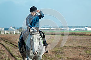 Little boy in a jockey cap on a white adult horse on a background of nature. Jockey, hippodrome, horseback riding