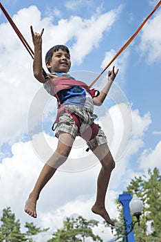 A little boy is jumping on a trampoline in the park. He soars in the air against the blue sky and shows a gesture that he is