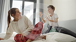 Little boy jumping from under the pile of cushions and scares mother sitting on bed. Concept of happy family, parents with kids,