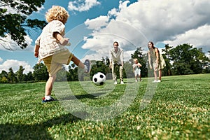 Little boy kicking the ball while parents and sister watching. Young family playing football on the grass field in the