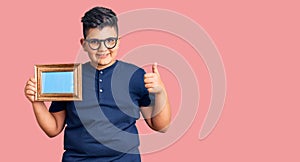 Little boy kid holding empty frame smiling happy and positive, thumb up doing excellent and approval sign