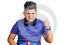 Little boy kid holding skate and wearing headphones annoyed and frustrated shouting with anger, yelling crazy with anger and hand