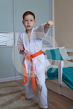 A little boy in a kimono with an orange belt stands in a fighting stance on a light background at home
