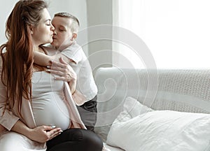 Little boy kisses and hugs his pregnant mom at home on the couch