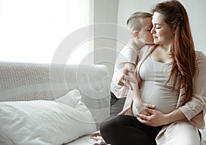 Little boy kisses and hugs his pregnant mom at home on the couch