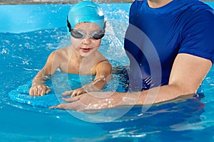 Little boy learning to swim in indoor pool with pool board and trainer