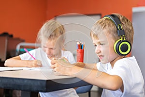 A little boy listening to music with headphones sitting at a table next to a girl student writing a text in a notebook