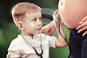 A little boy listens with interest to the stomach of a pregnant mother with a stethoscope.