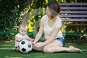 Little boy looking at soccer ball and exploring it sitting next to mother at football field. Toddler son playing with mom outdoors