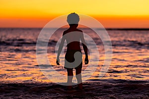 Little boy looking at sunset at the ocean and preparing to swim