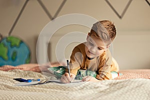 Little boy lying on a bed and doing his homework.