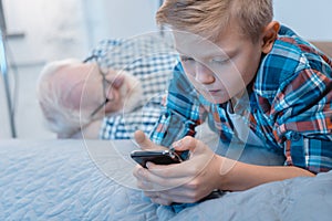 Little boy lying on bed and using his smartphone while his granpa is napping