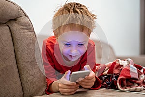 Little boy is lying on couch with smartphone in hands and playing mobile game or using social networks. Modern problem