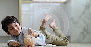 Little boy lying on floor indoor putting coins into piggybank