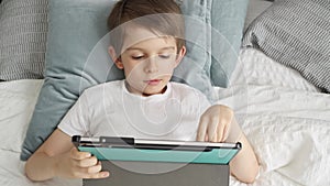 A little boy is lying on his bed and playing with a tablet computer during the day. Child using gadget, education.