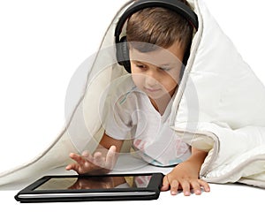 Little boy lying under blanket with tablet in headphones