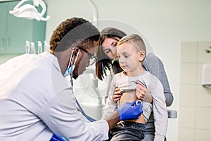 Little boy with mother visiting children`s African American doctor with stethoscope in hospital