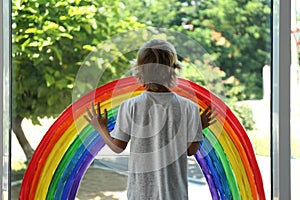 Little boy near rainbow painting on window indoors. Stay at home concept