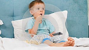 Little boy in pajamas eating popcorn in bed at morning and switching cartoons on TV with remote control