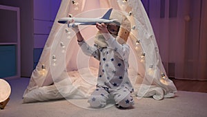 Little boy in pajamas, glasses and an aviapilot hat is sitting in dark room with white handmade tent, wigwam with
