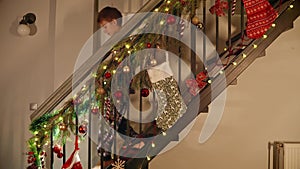 Little boy in pajamas at night walking down the wooden stairs at house decorated for Christmas or New Year celebrations