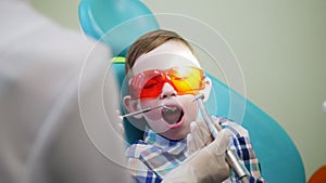 Little boy patient in yellow glasses sitting in a chair. At this time, the dentist treats his teeth. The patient`s