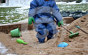 Little boy playing in blue winter overalls and green fingerless gloves on the sandpit. He holds habits and digs sand. There is a w