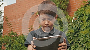 Little boy playing games on his touchscreen tablet outdoors in front of the house covered in greenery