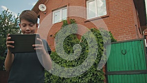 Little boy playing games on his touchscreen tablet outdoors in front of the house covered in greenery