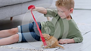 Little boy playing with ginger kitten red ribbon at home on the floor. Friendship of children and animals, friends and