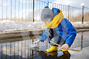 Little boy playing with origami paper boat in large puddle near town houses. Paper ship floating in water