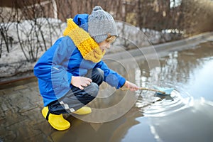 Little boy playing with paper boat in large puddle near town houses in early spring. Paper ship floating in water