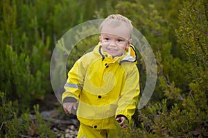 Little boy playing in rainy summer park. Child with colorful rainbow umbrella, waterproof coat and boots jumping in