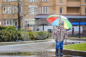 Little boy playing in rainy summer park. Child with colorful rainbow umbrella, waterproof coat and boots jumping in puddle and mu