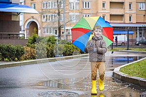 Little boy playing in rainy summer park. Child with colorful rainbow umbrella, waterproof coat and boots jumping in puddle and mu