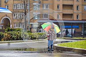 Little boy playing in rainy summer park. Child with colorful rainbow umbrella, waterproof coat and boots jumping in puddle and mu