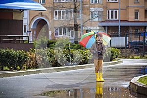 Little boy playing in rainy summer park. Child with colorful rainbow umbrella, waterproof coat and boots jumping in puddle and mu